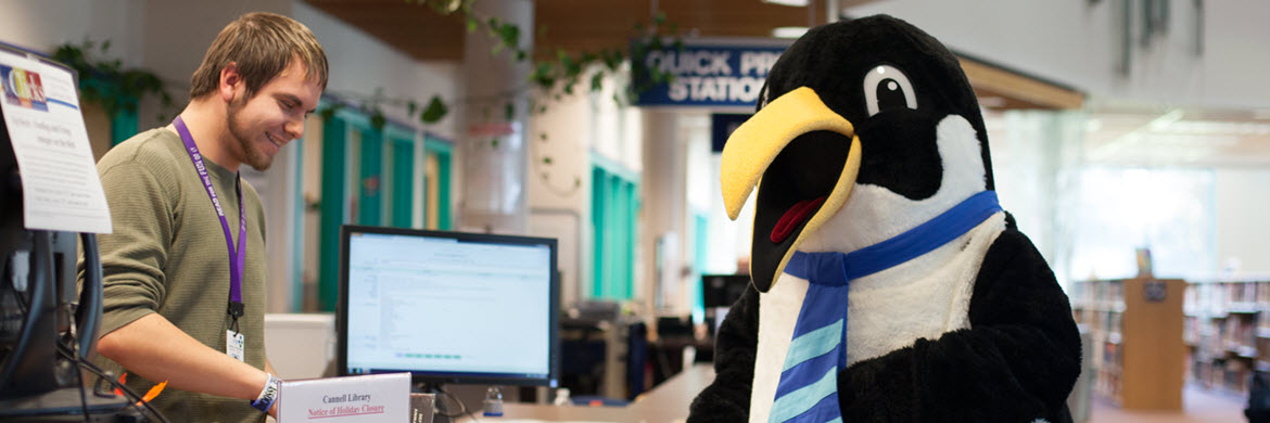 Oswald the penguin mascot in front of Check Out desk with smiling young man behind the counter